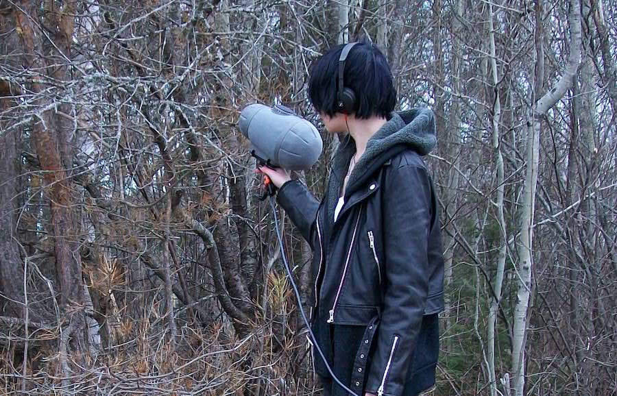 Woman in leather jacket pointing a field recording microphone towards a mixed forest.